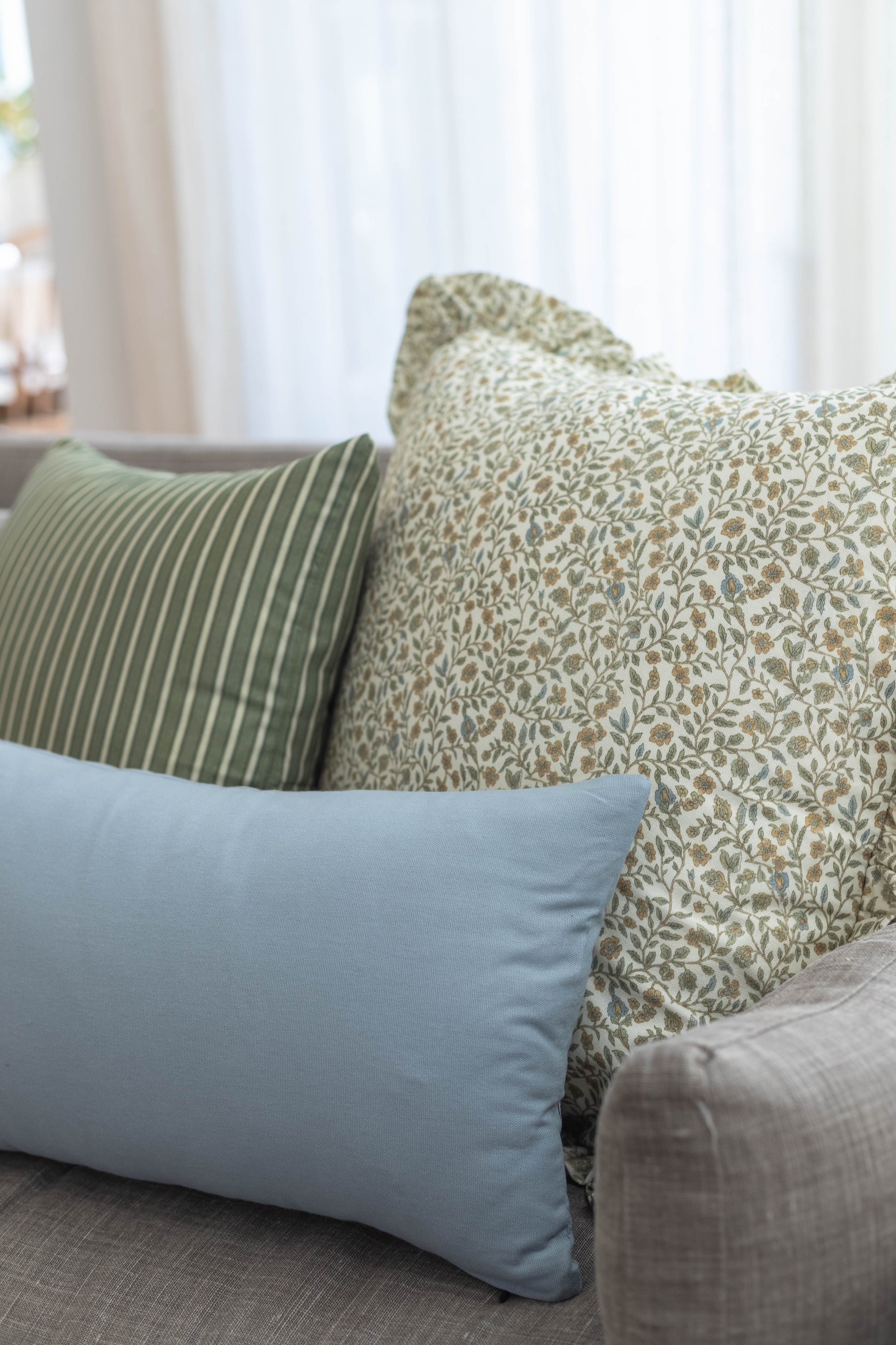 Decorative pillows in sage green striped, cream floral, and blue solid patterns arranged on a sofa.