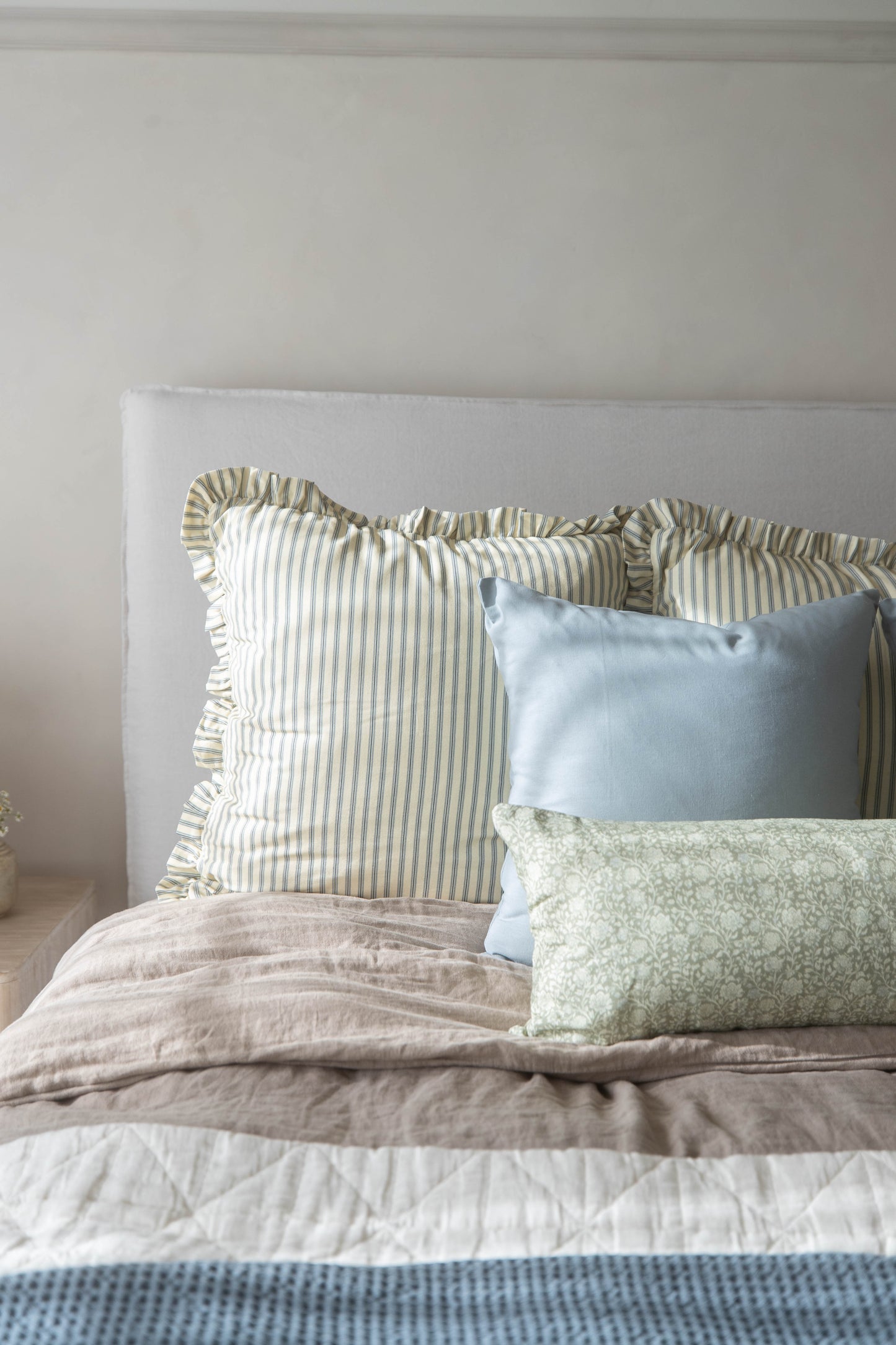 Striped linen pillow with ruffled edges in sage and cream, styled with solid blue and patterned pillows on a bed.