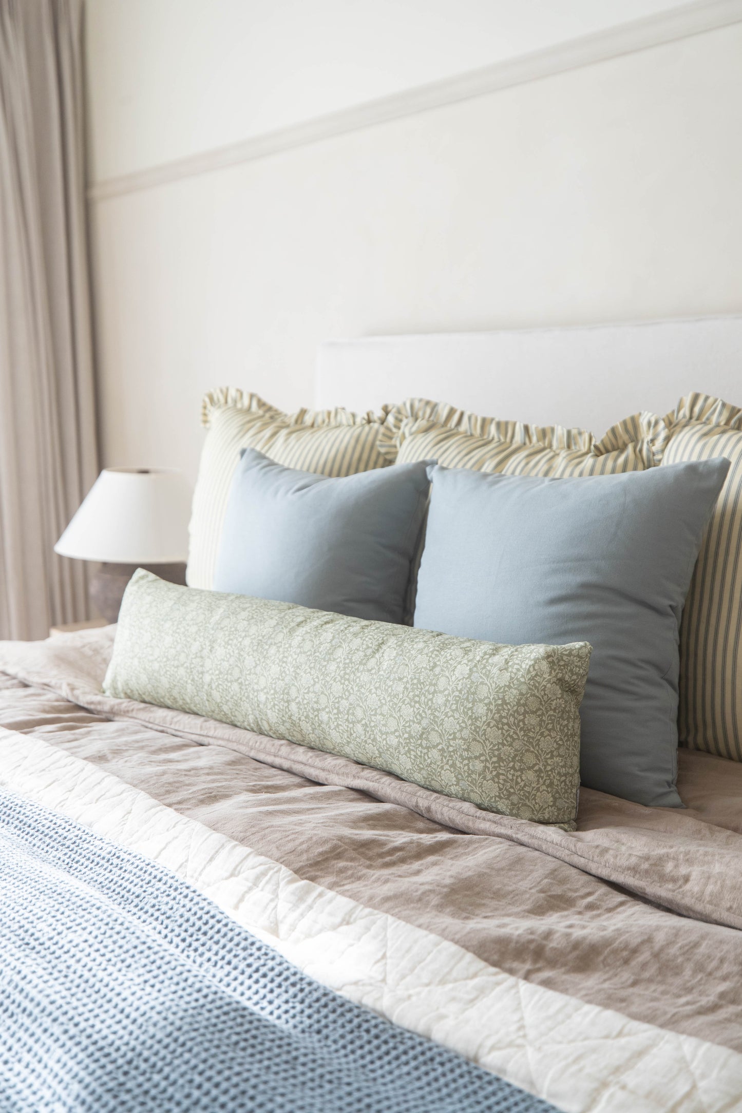 Bed with blue and striped pillows, green patterned lumbar pillow, beige bedding, white headboard, and neutral curtains.