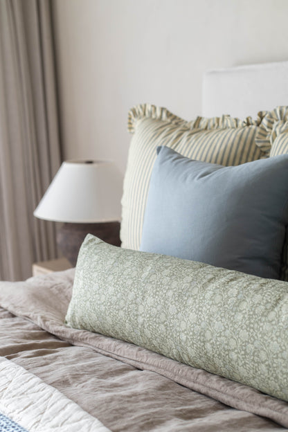 Bed with green patterned pillow, blue pillow, and striped ruffled pillows, with beige bedding and nightstand lamp.