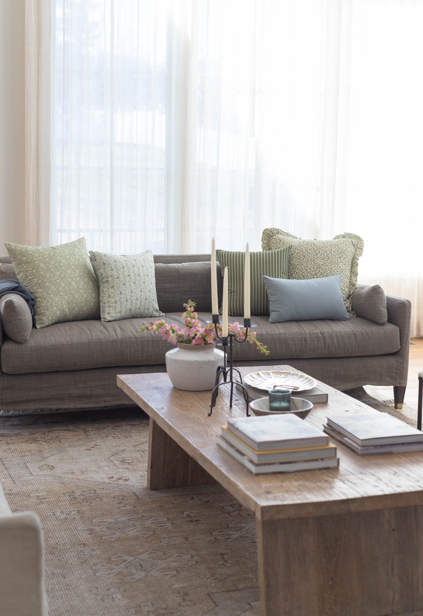 Green and blue patterned throw pillow with ruffled edges displayed on gray sectional sofa in bright living room.