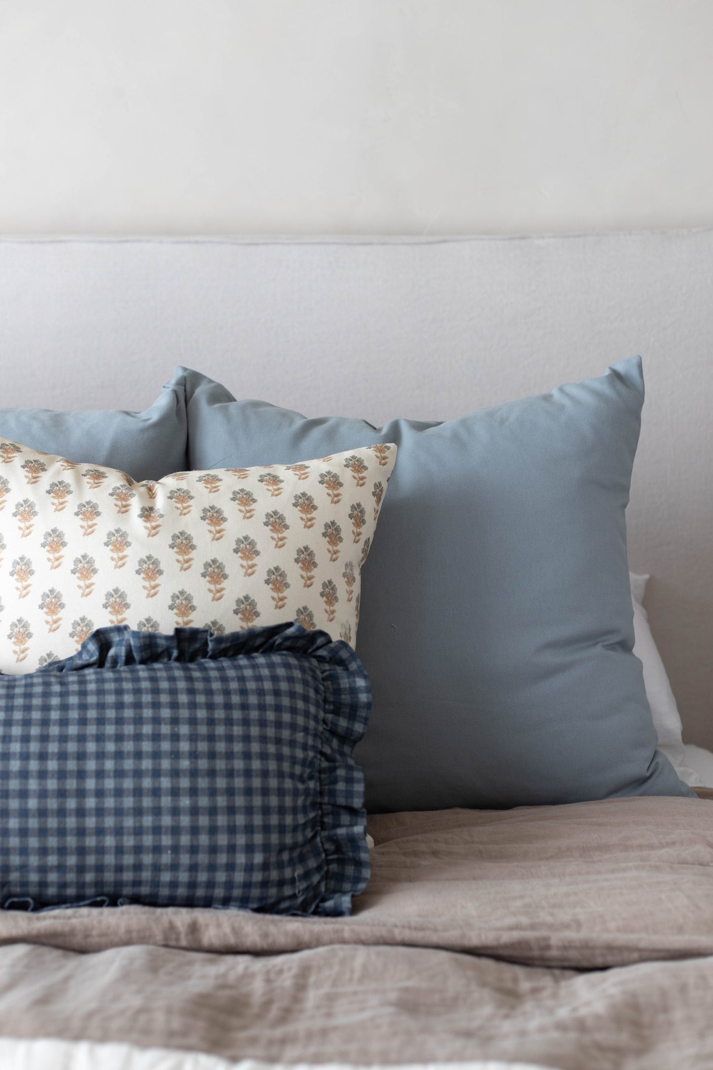 Three decorative pillows arranged on a bed: a floral patterned pillow, a blue-gray solid pillow, and a navy gingham pillow wi