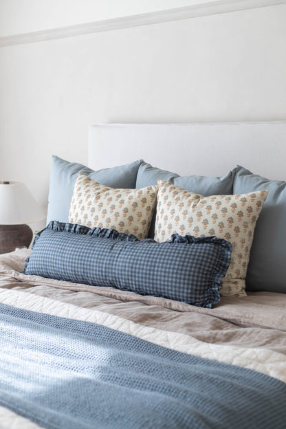 Bed with white headboard, gray and blue pillows, beige floral pillows, and blue gingham bolster pillow.