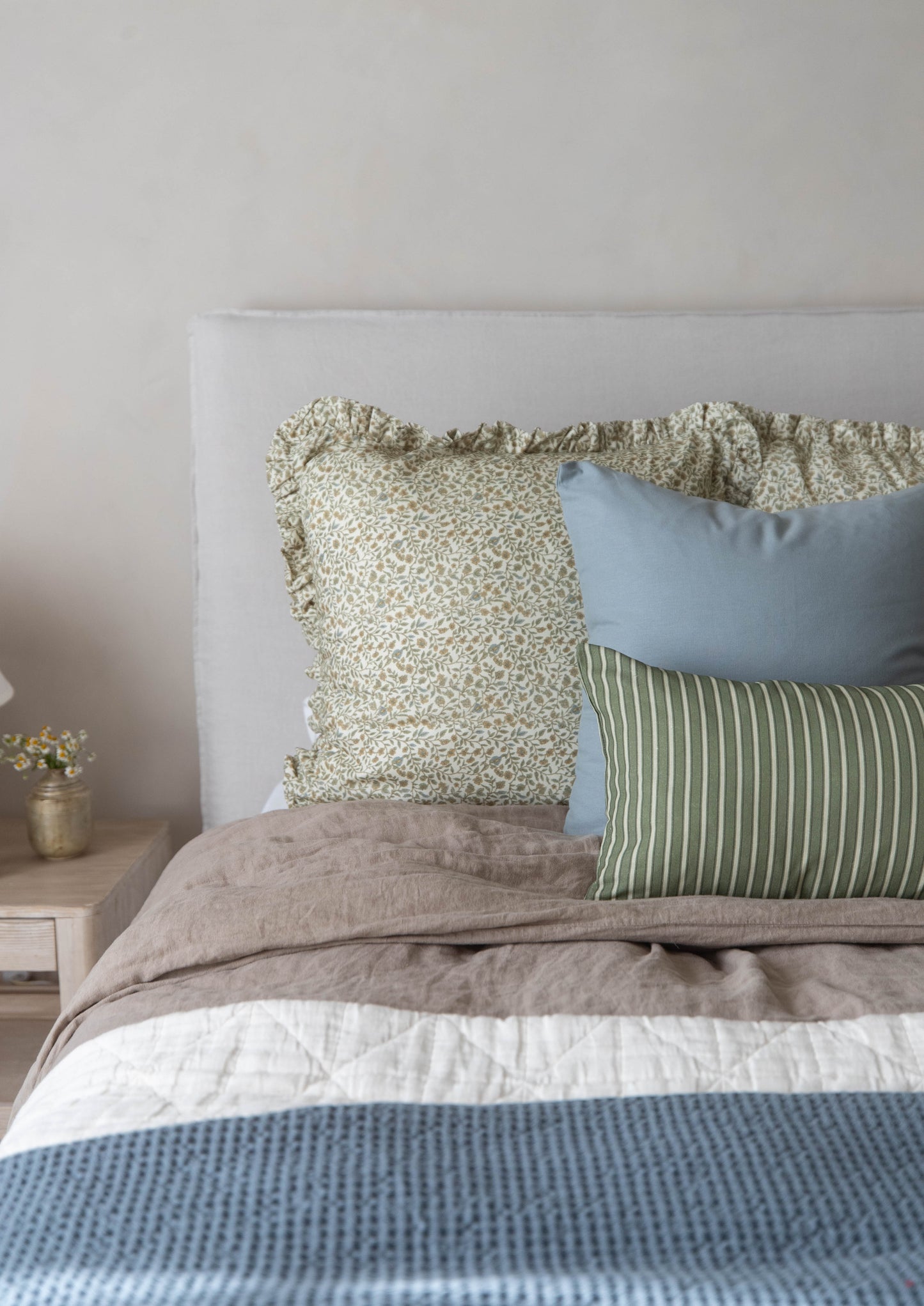 Bed styled with layered pillows in sage floral, blue, and green stripe patterns, tan textured bedding, and blue waffle-weave 