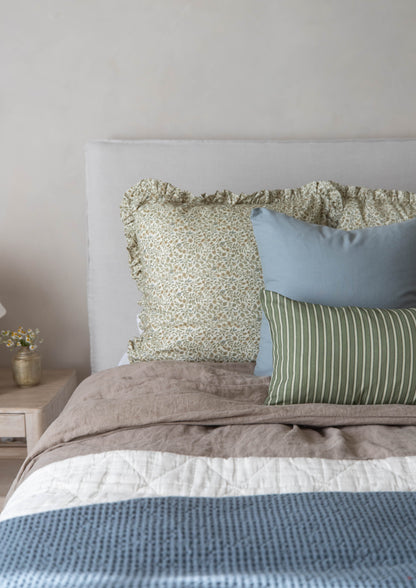 Bed styled with layered pillows in sage floral, blue, and green stripe patterns, tan textured bedding, and blue waffle-weave 