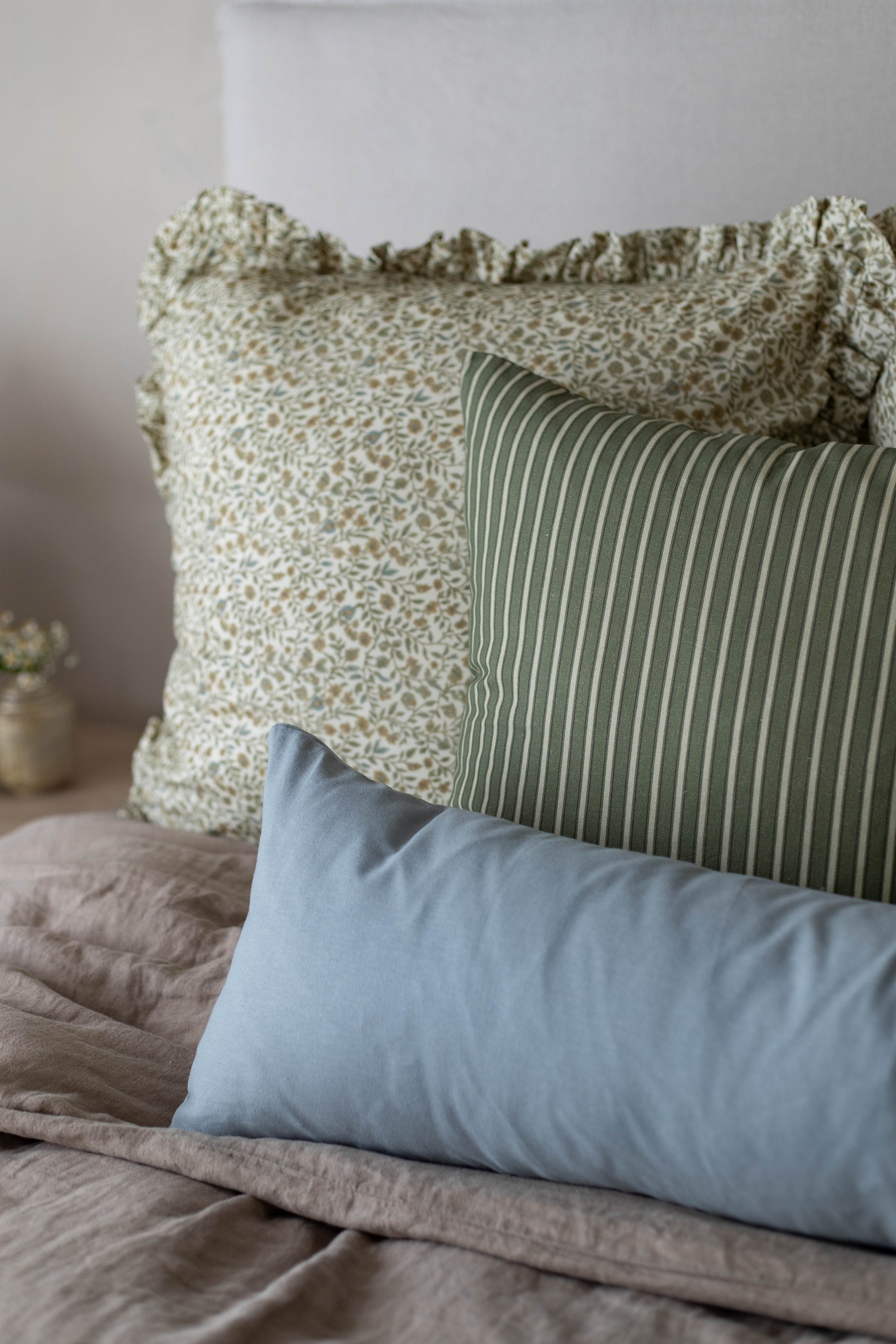 Three decorative pillows with ruffled edges in cream textured, green striped, and blue solid fabrics arranged on a bed.