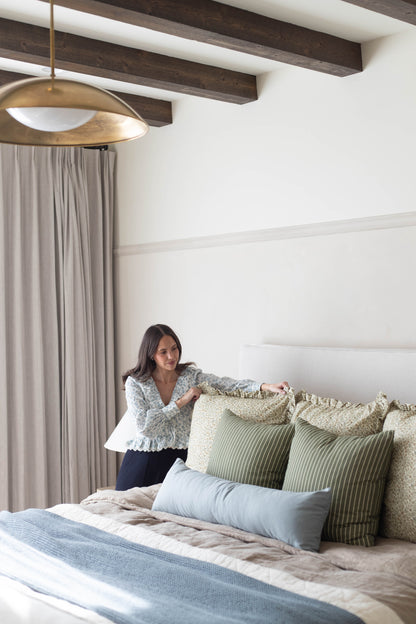 Brass pendant light with white interior hanging above a bed with decorative pillows and headboard.