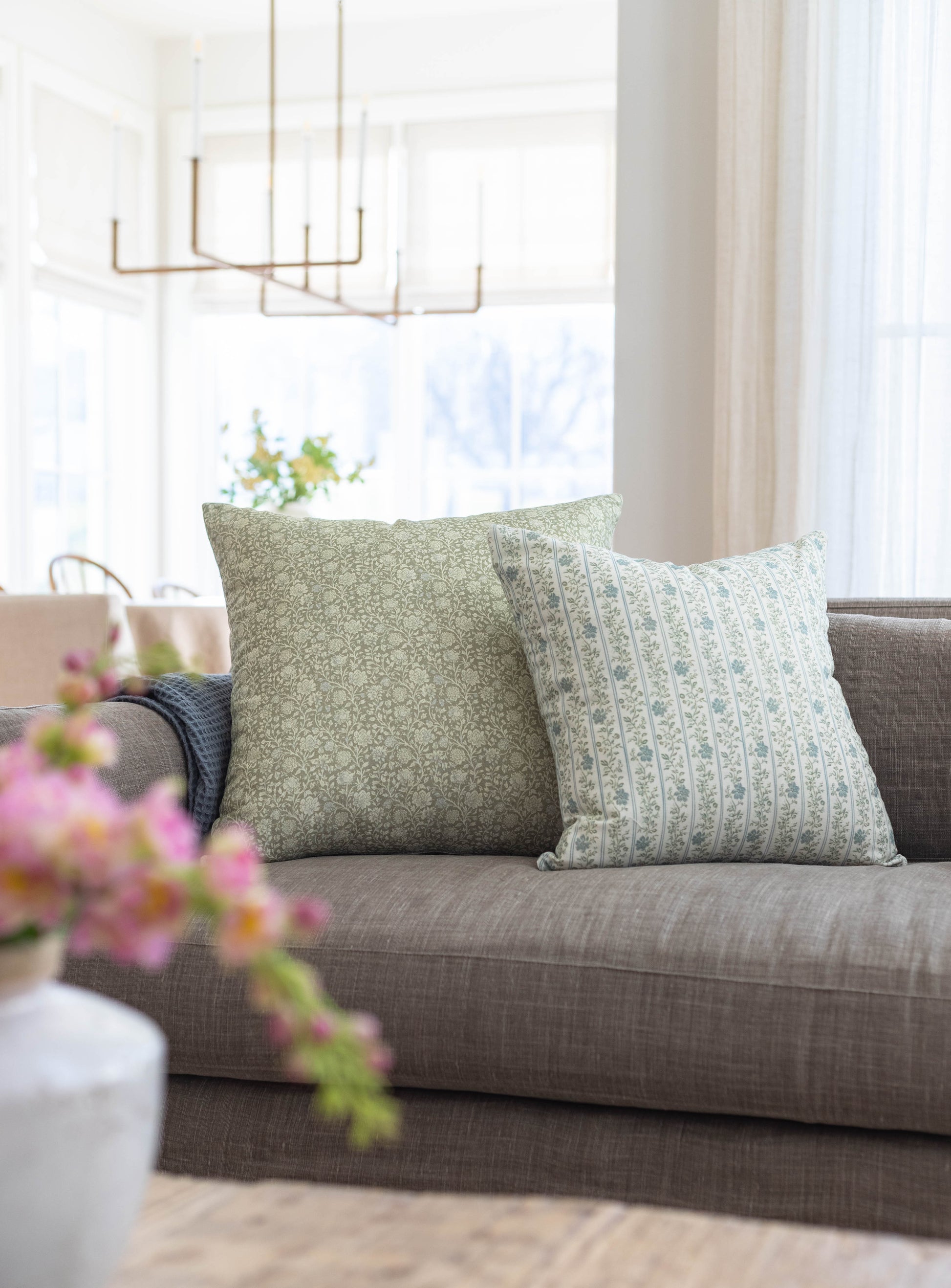 Two decorative pillows with sage green floral patterns on a gray upholstered sofa near a window.