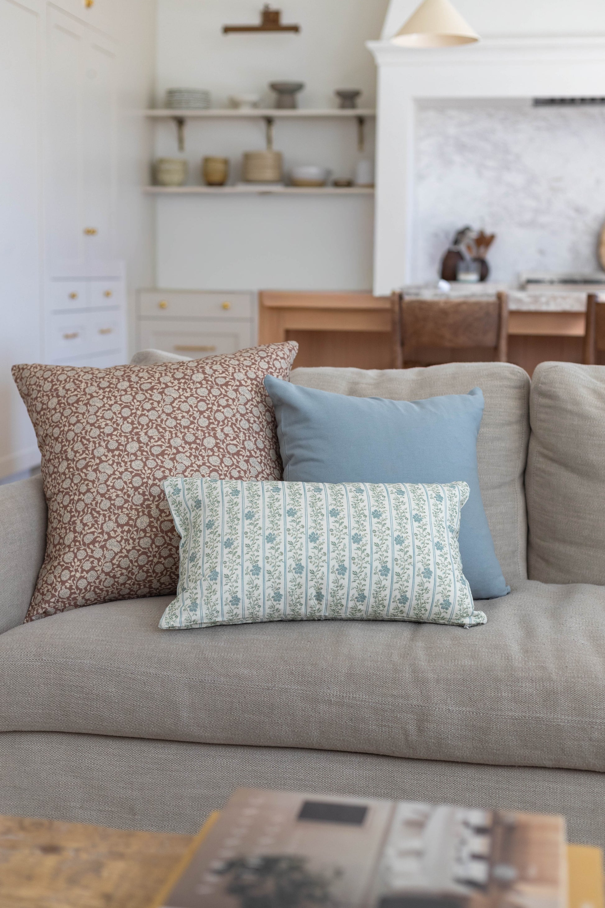 Three decorative pillows on a beige sofa: brown floral, light blue solid, and cream striped with blue pattern.