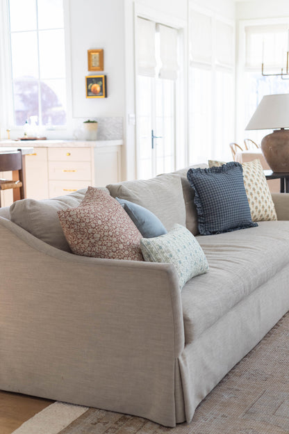 Beige upholstered sofa with four decorative pillows in various patterns positioned in a bright living room with kitchen and d