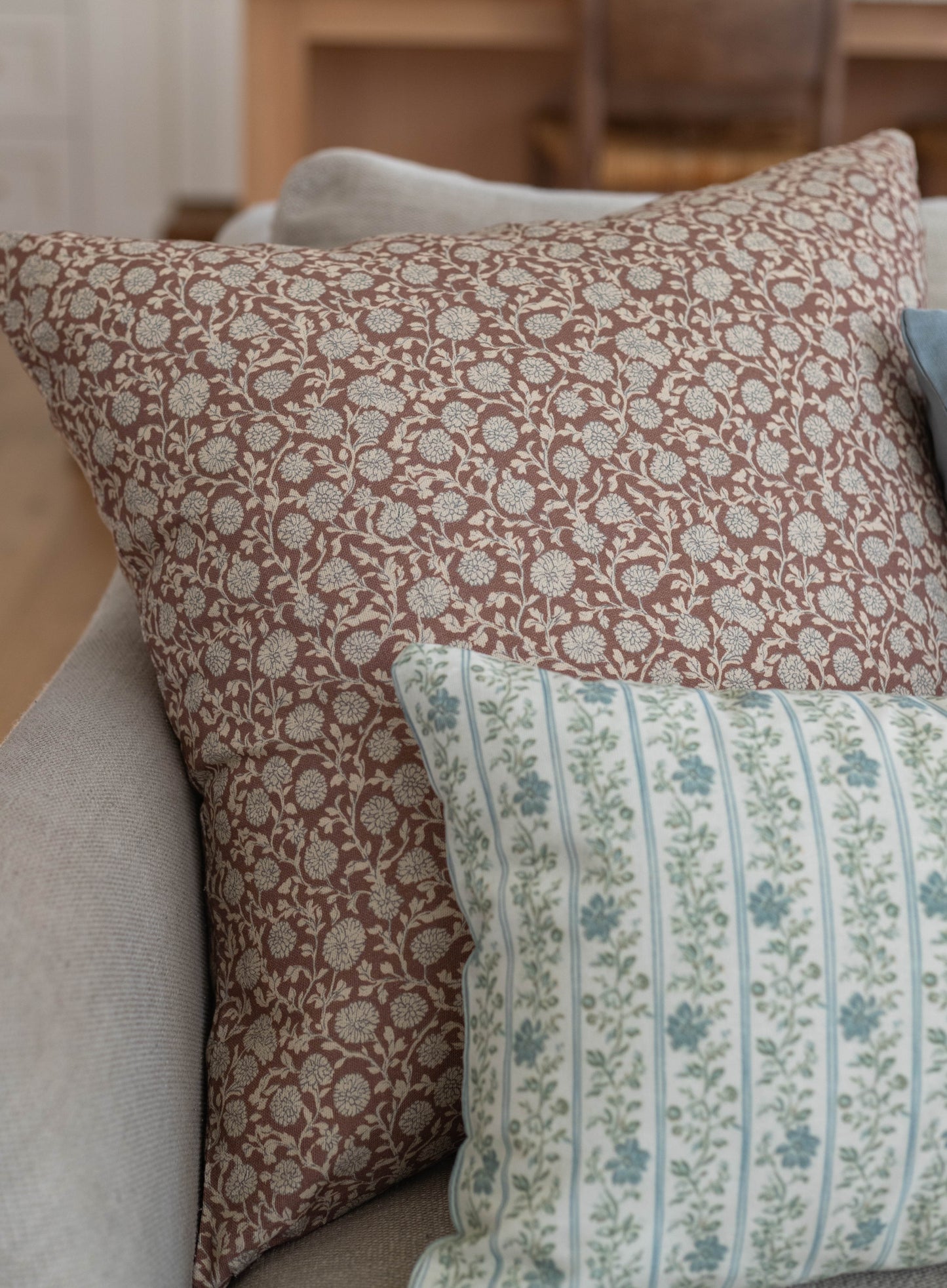 Tan and cream floral patterned pillow with dense leaf and botanical motifs displayed on a neutral upholstered surface.