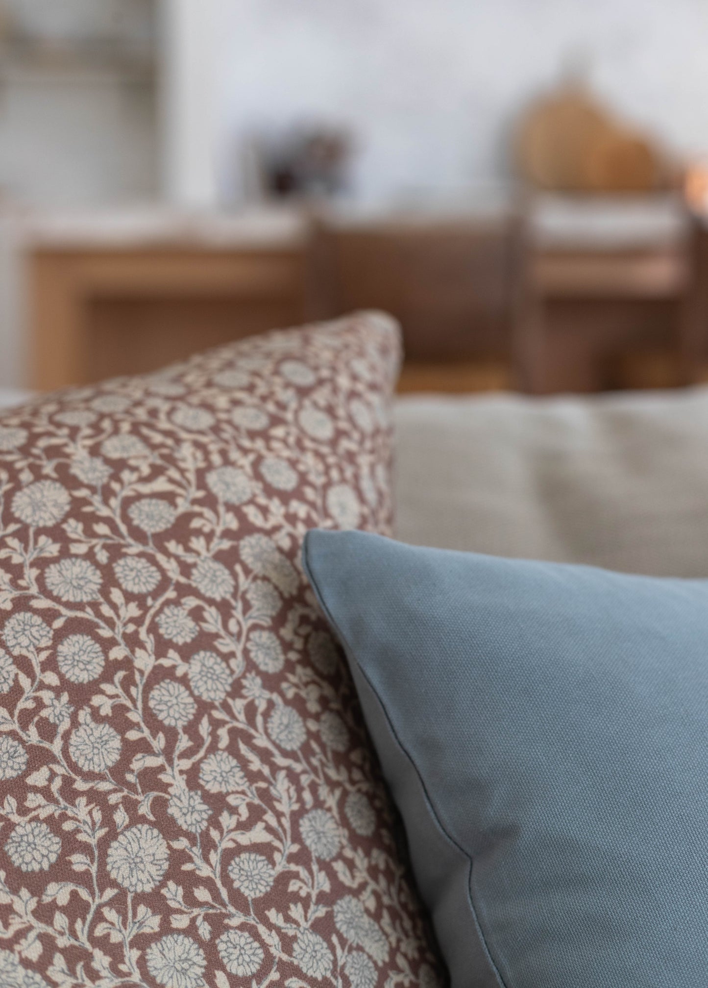 Decorative pillow with floral pattern in taupe and cream displayed on a light blue pillow against a blurred interior backgrou