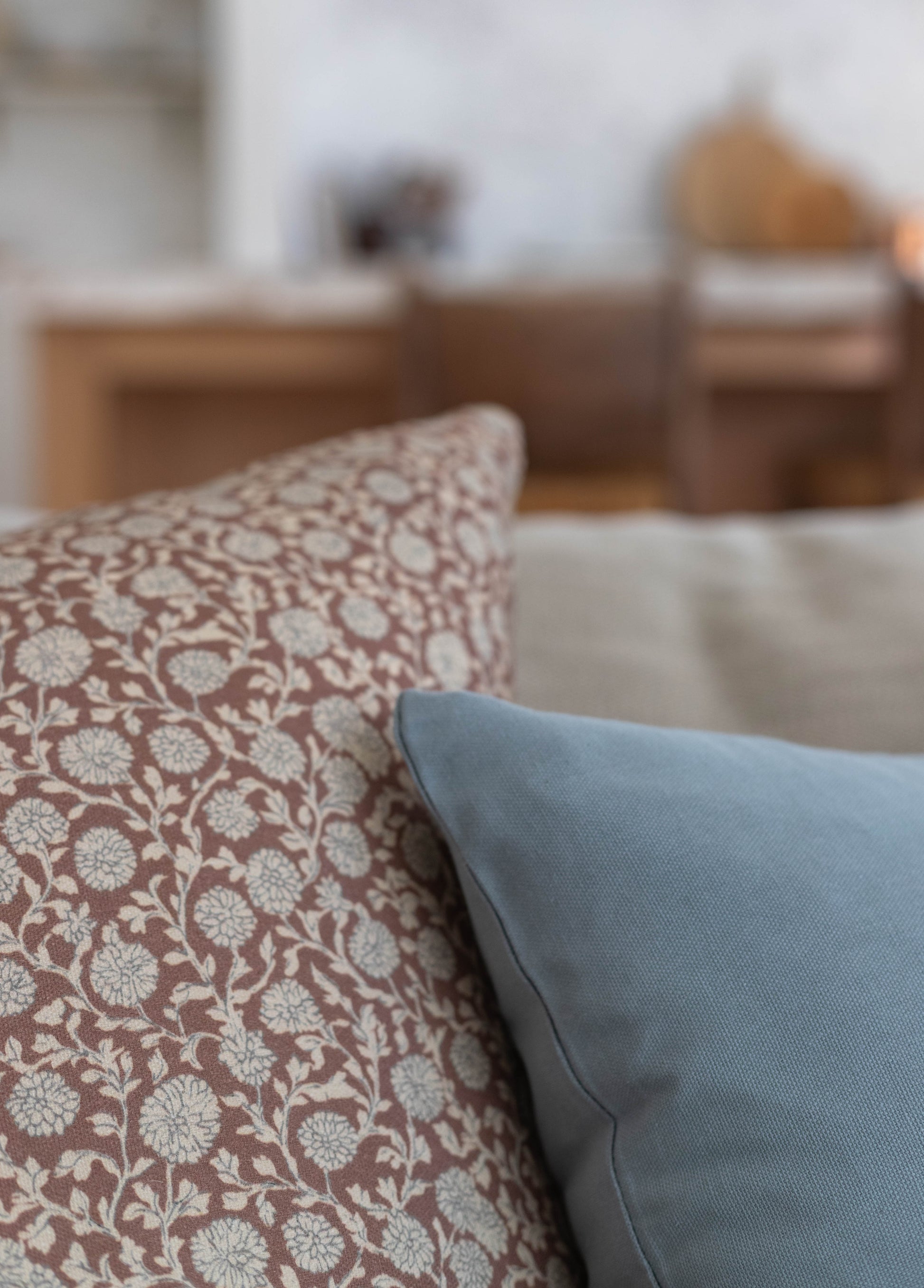 Decorative pillow with floral pattern in taupe and cream displayed on a light blue pillow against a blurred interior backgrou
