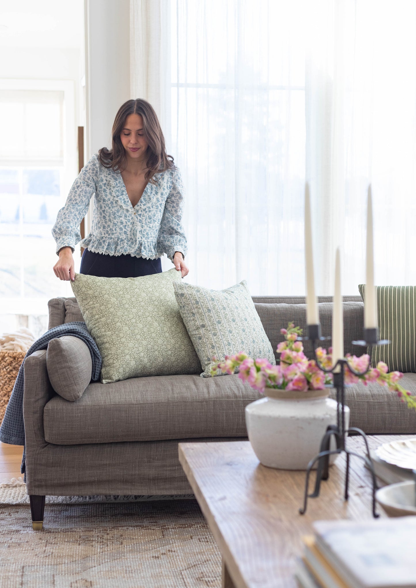 Woman holding two green patterned throw pillows on a gray upholstered sofa in a bright living room.