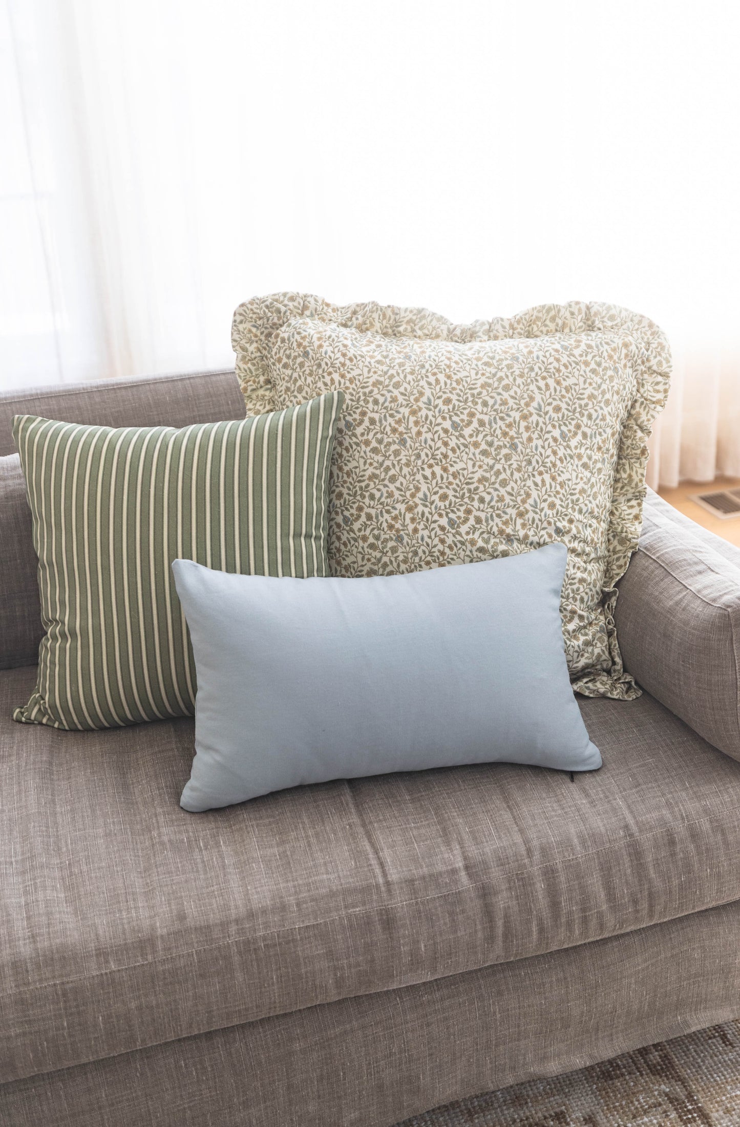 Three decorative pillows on a gray sofa: green striped, beige textured with ruffle trim, and light blue solid.