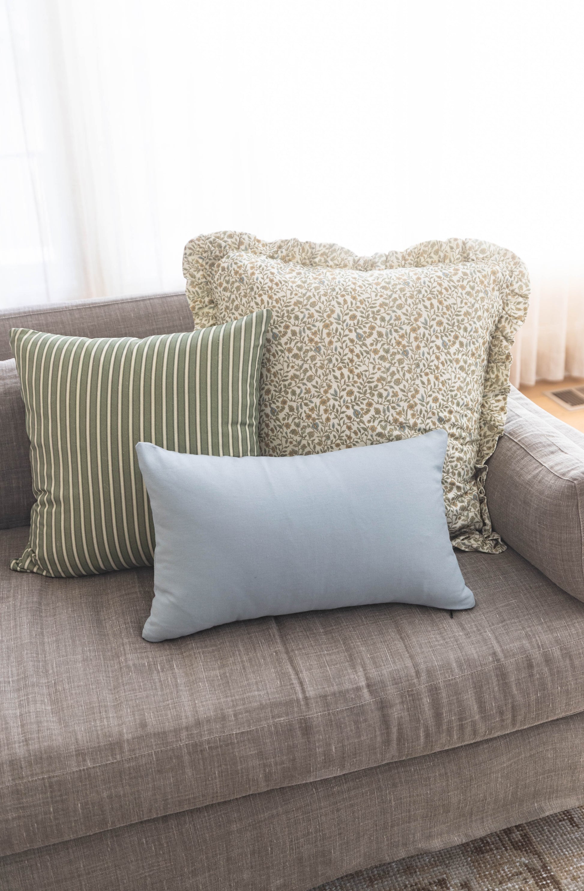 Three decorative pillows on a gray sofa: green striped, beige textured with ruffle trim, and light blue solid.