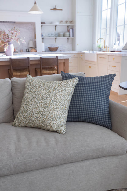 Two decorative pillows on a gray sofa: one with beige leaf pattern, one with dark blue gingham check.