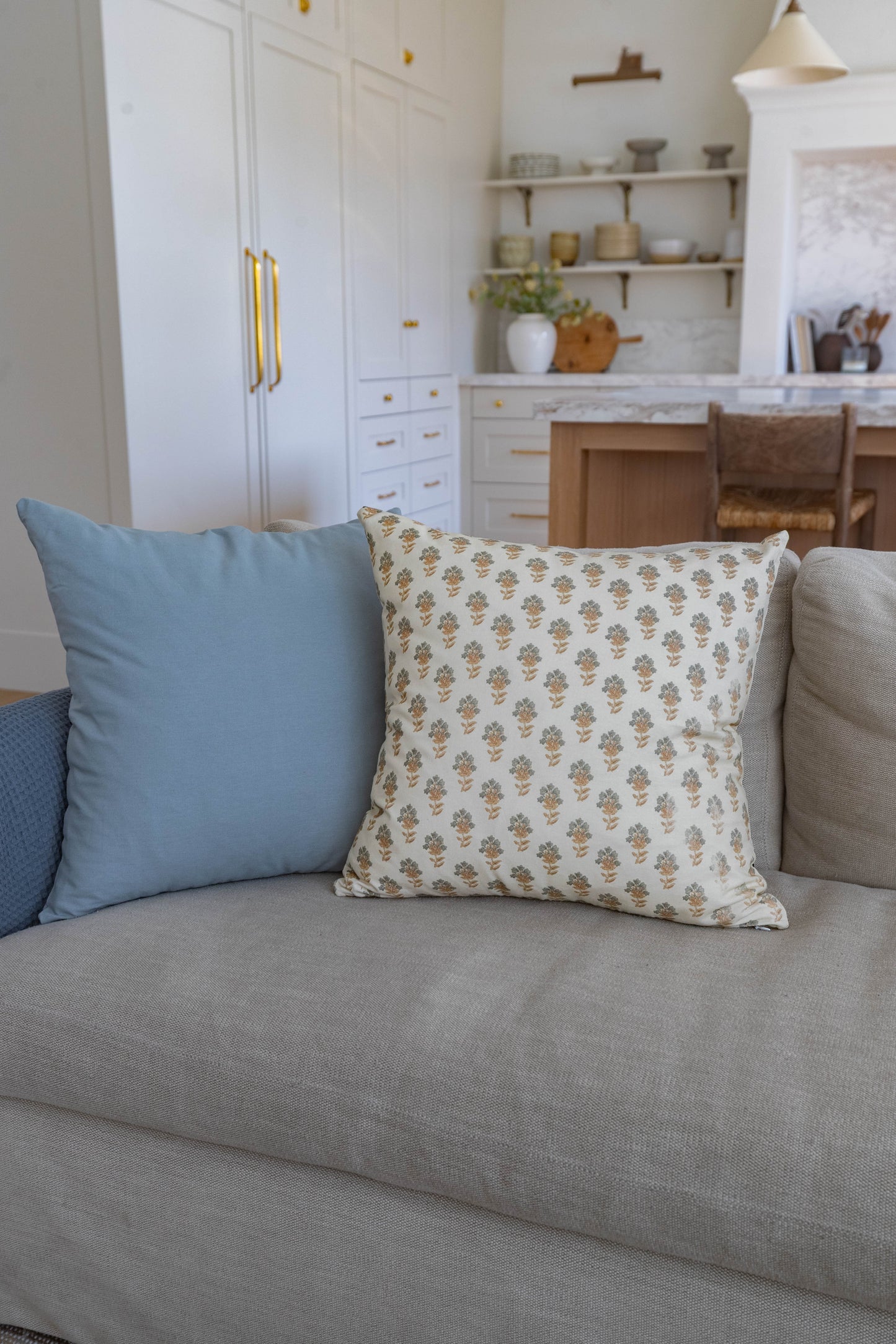 Cream throw pillow with gold leaf print displayed on light gray sofa with blue pillow.