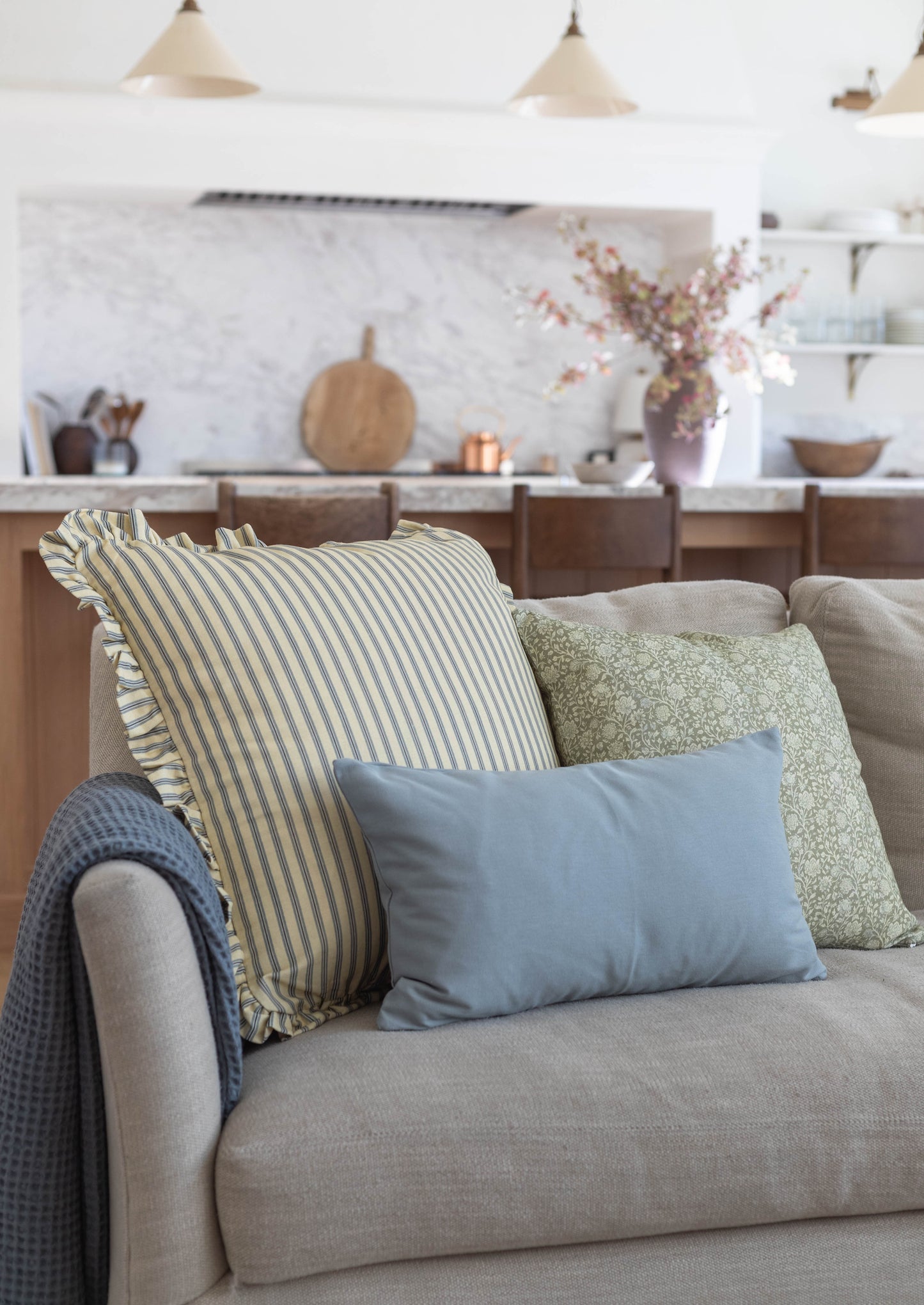 Striped cream and blue pillow with ruffled edges displayed on a beige sofa with coordinating throw pillows.