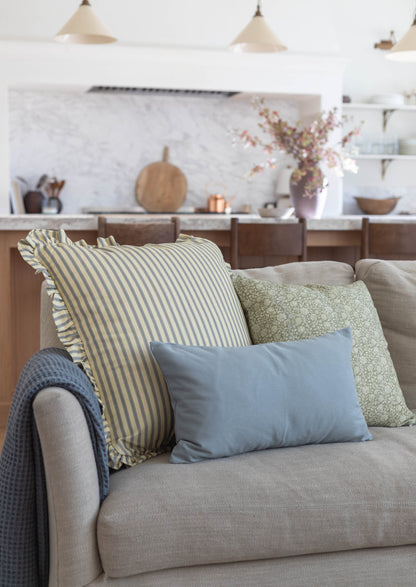 Striped cream and blue pillow with ruffled edges displayed on a beige sofa with coordinating throw pillows.
