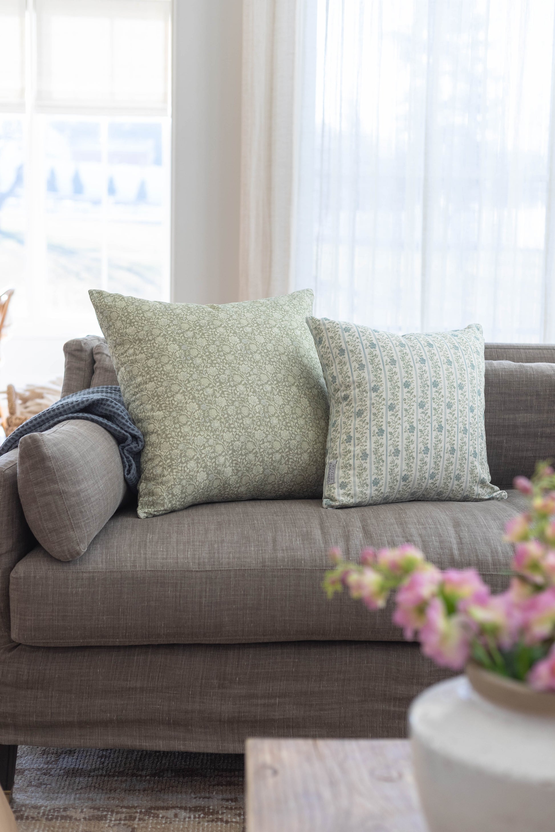 Gray upholstered sofa with green and blue patterned throw pillows near a window.