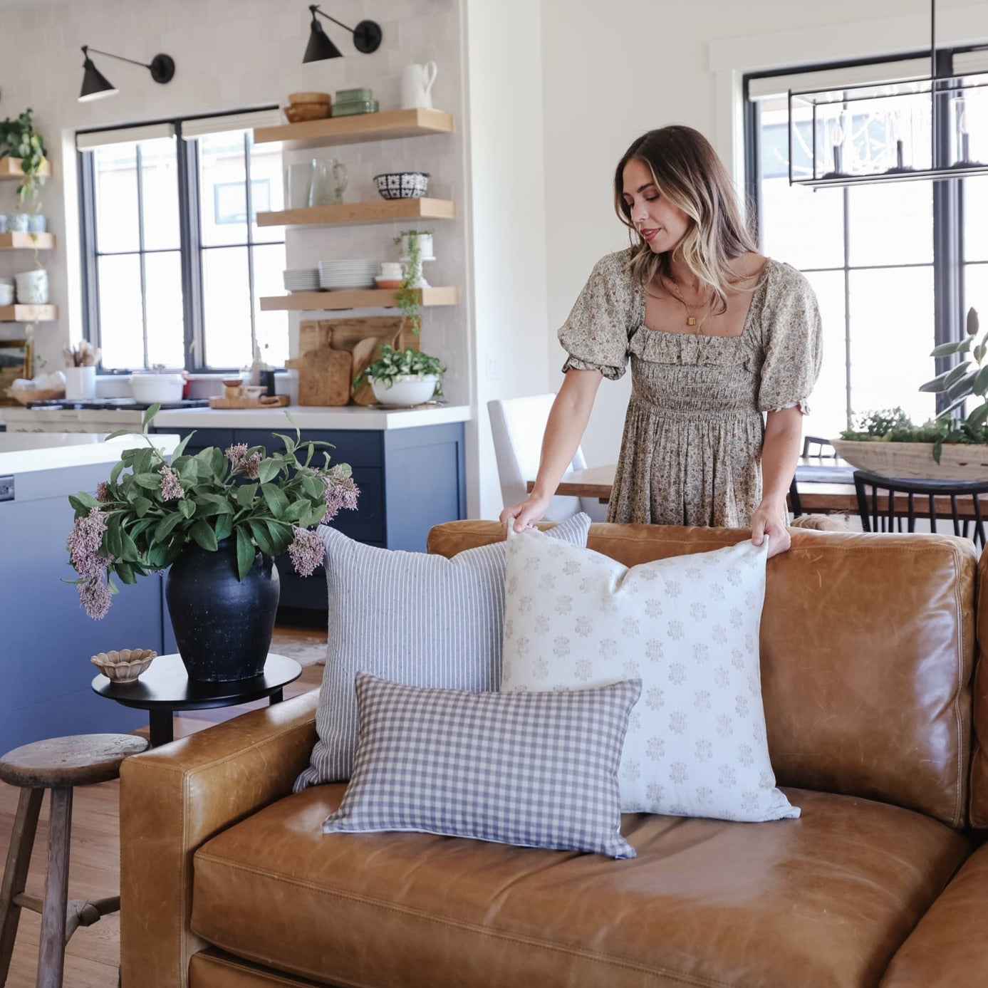Woman arranging patterned throw pillows on a tan leather sofa in a modern kitchen and living room.