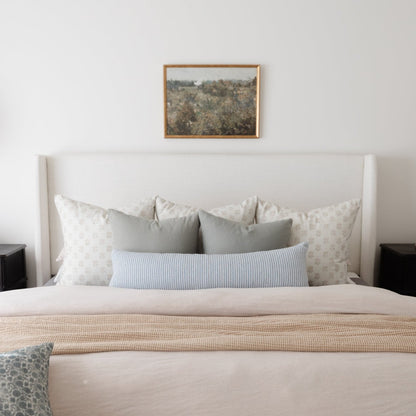 Framed landscape artwork with natural wood border hanging above a bed with patterned pillows.