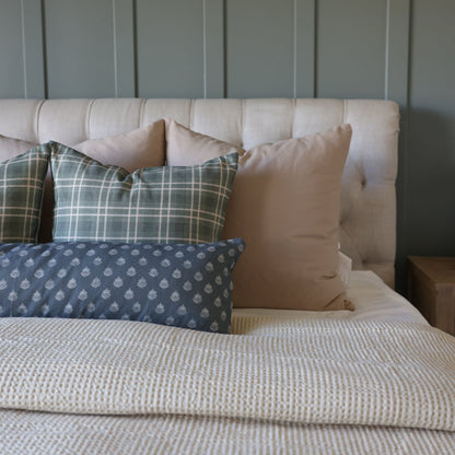 Bed styled with four decorative pillows in plaid, solid tan, and patterned blue designs against a cream upholstered headboard
