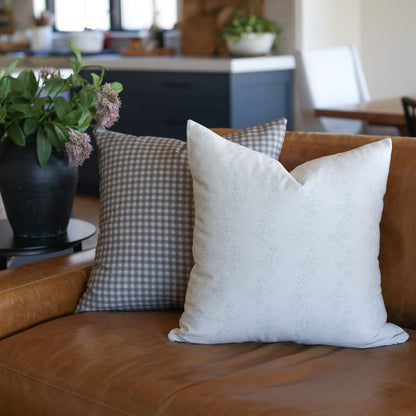 Cream throw pillow with subtle texture pattern displayed on tan leather sofa beside gray gingham pillow.