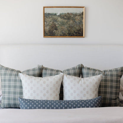 Framed landscape print with tan wood border hanging above bed with patterned pillows.