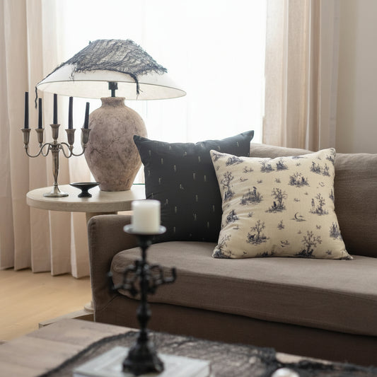 A styled living room scene featuring a gray sofa with two decorative throw pillows, one navy with skeletons and and one cream with a dark blue toile de Jouy pattern. 