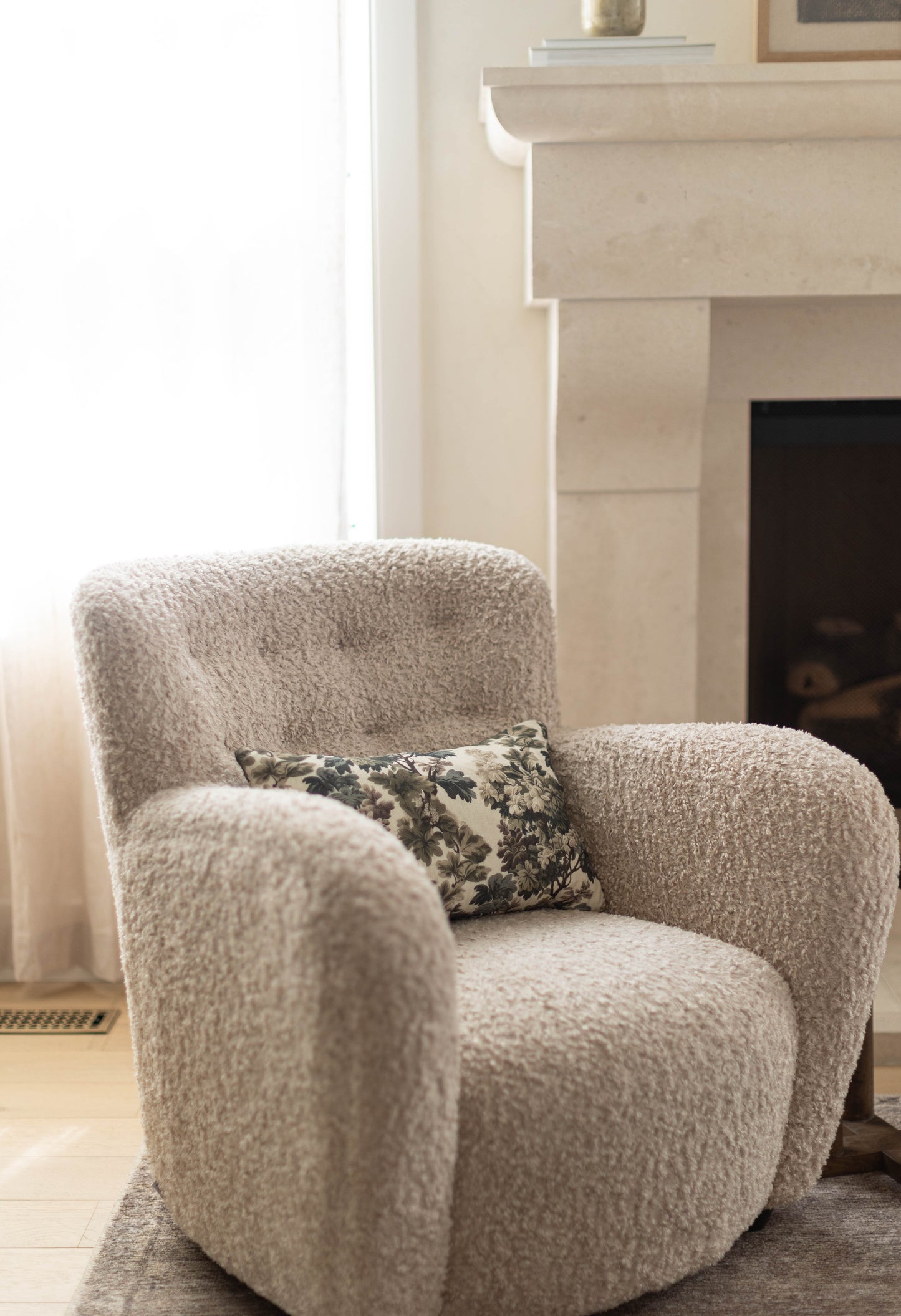 Beige textured armchair with curved arms and back, featuring a small black and cream floral pillow on the seat.
