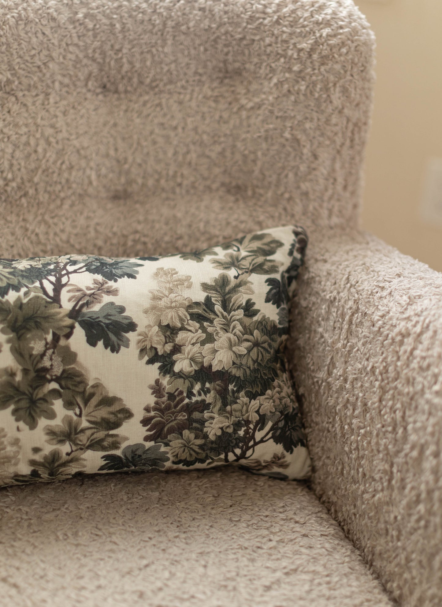 Beige textured pillow with floral botanical print in cream, green, and brown tones resting on upholstered furniture.