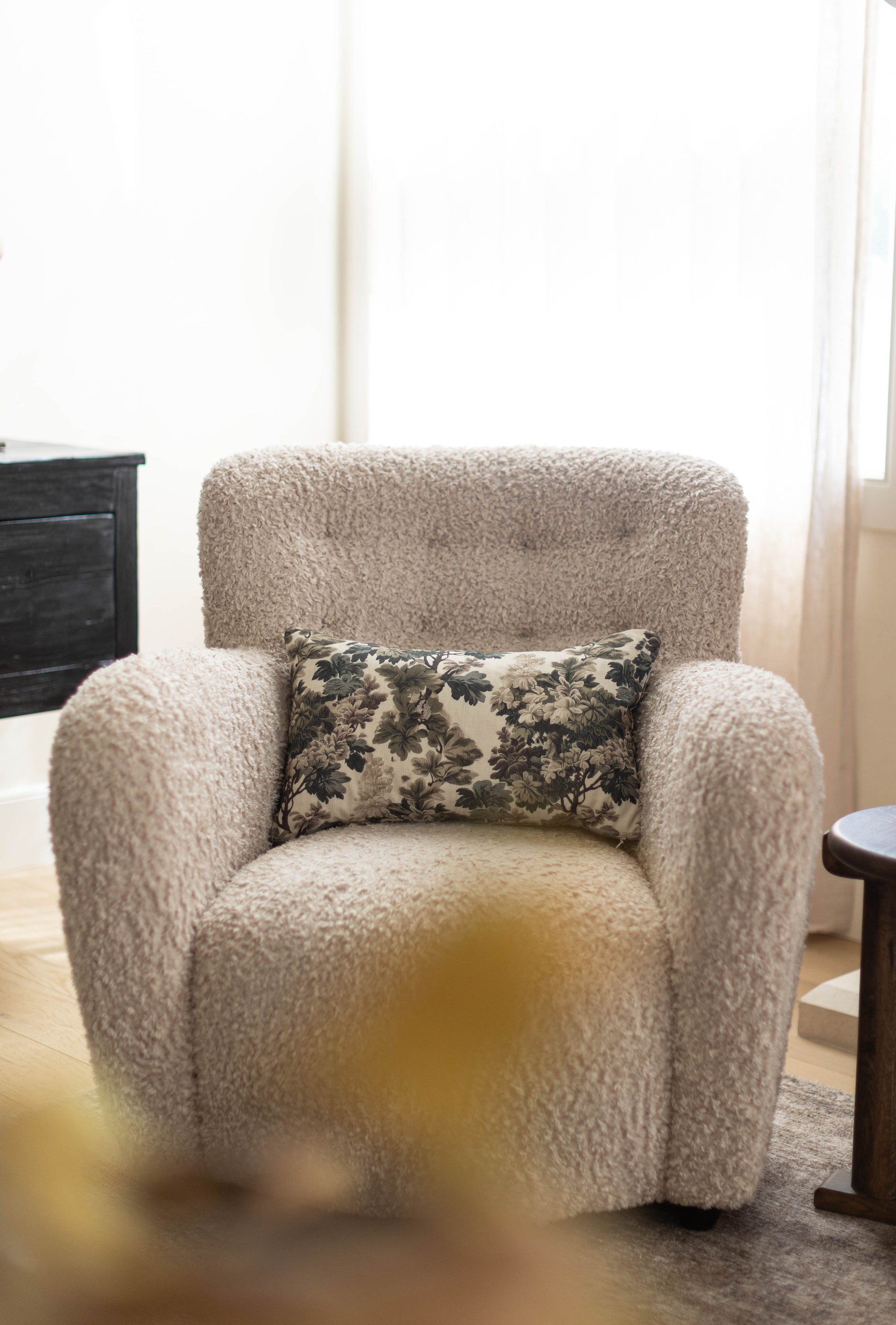 Beige textured armchair with rounded arms and back, featuring a black and cream floral rectangular pillow.