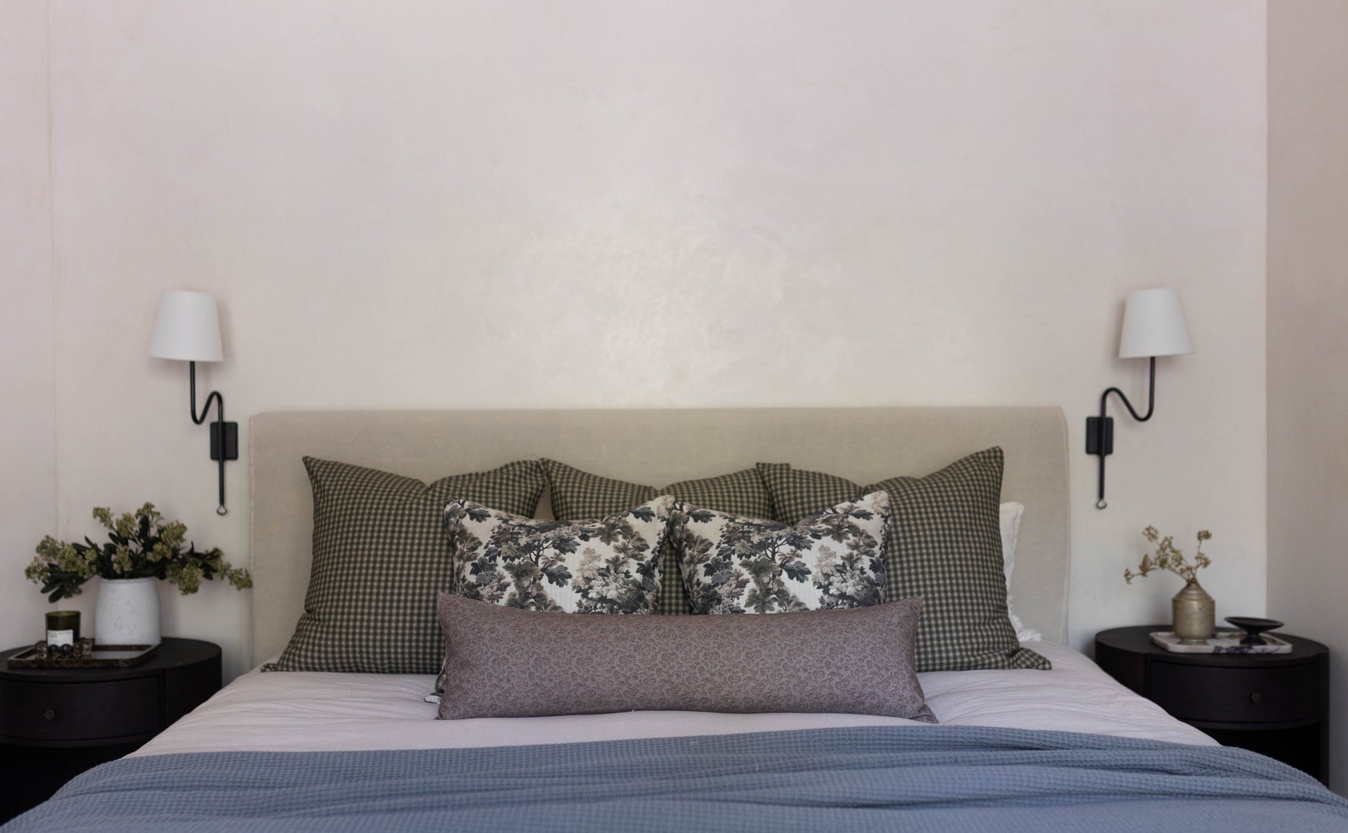 Bed with tan headboard dressed with gray checkered pillows, floral and gray throw pillows, and blue bedding, flanked by black