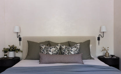 Bed with tan headboard dressed with gray checkered pillows, floral and gray throw pillows, and blue bedding, flanked by black