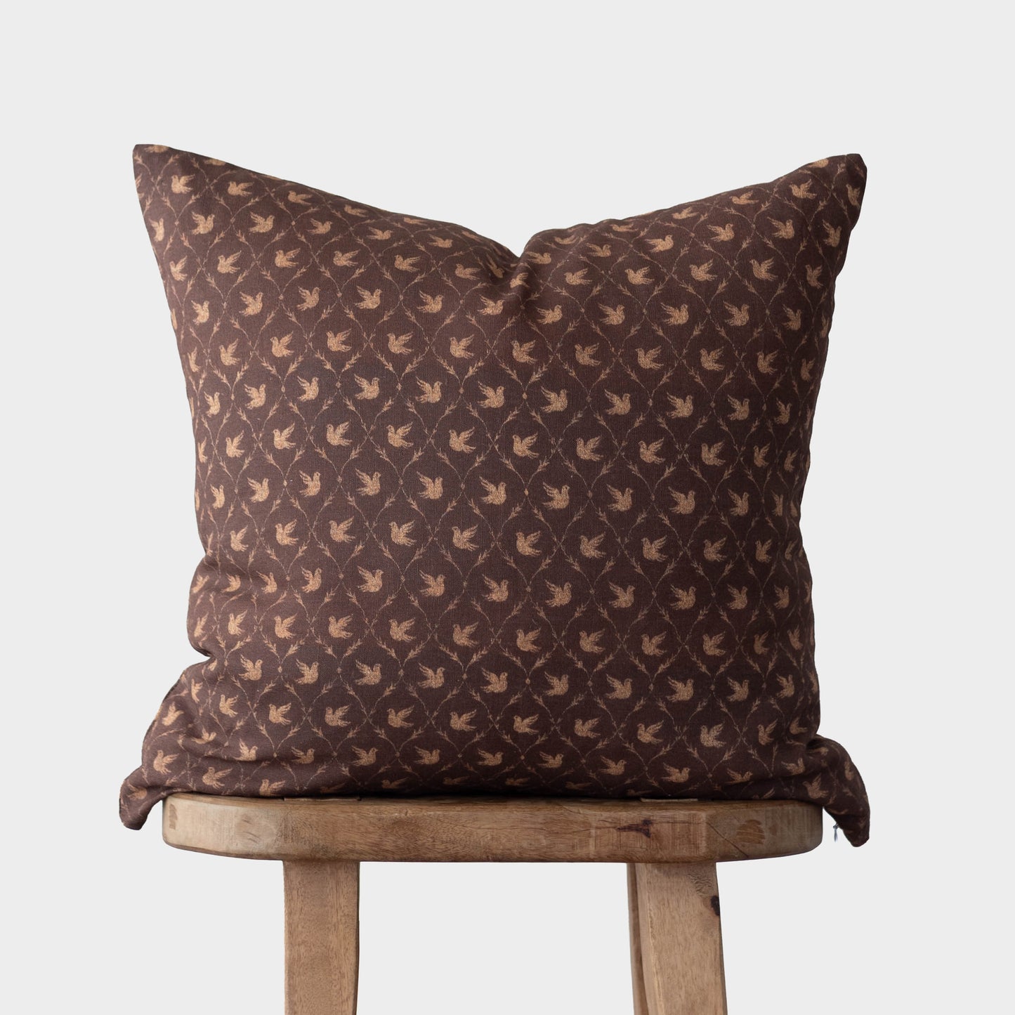 Brown cushion cover with repeating dove pattern in tan, displayed on wooden stool.
