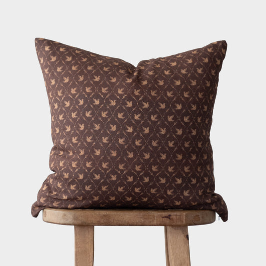 Brown cushion cover with repeating dove pattern in tan, displayed on wooden stool.