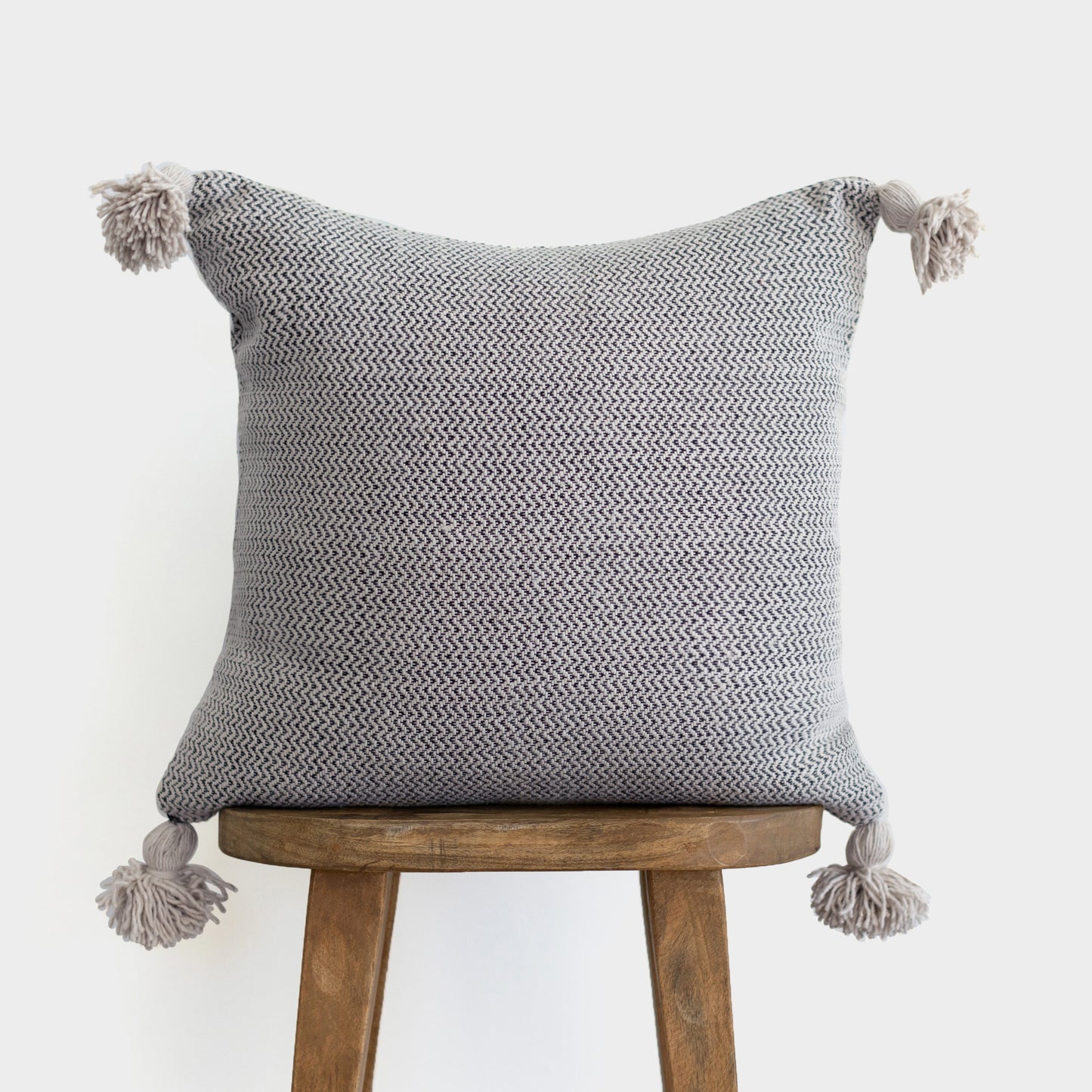 Gray tweed pillow with chevron pattern and tassel corners displayed on wooden stool.