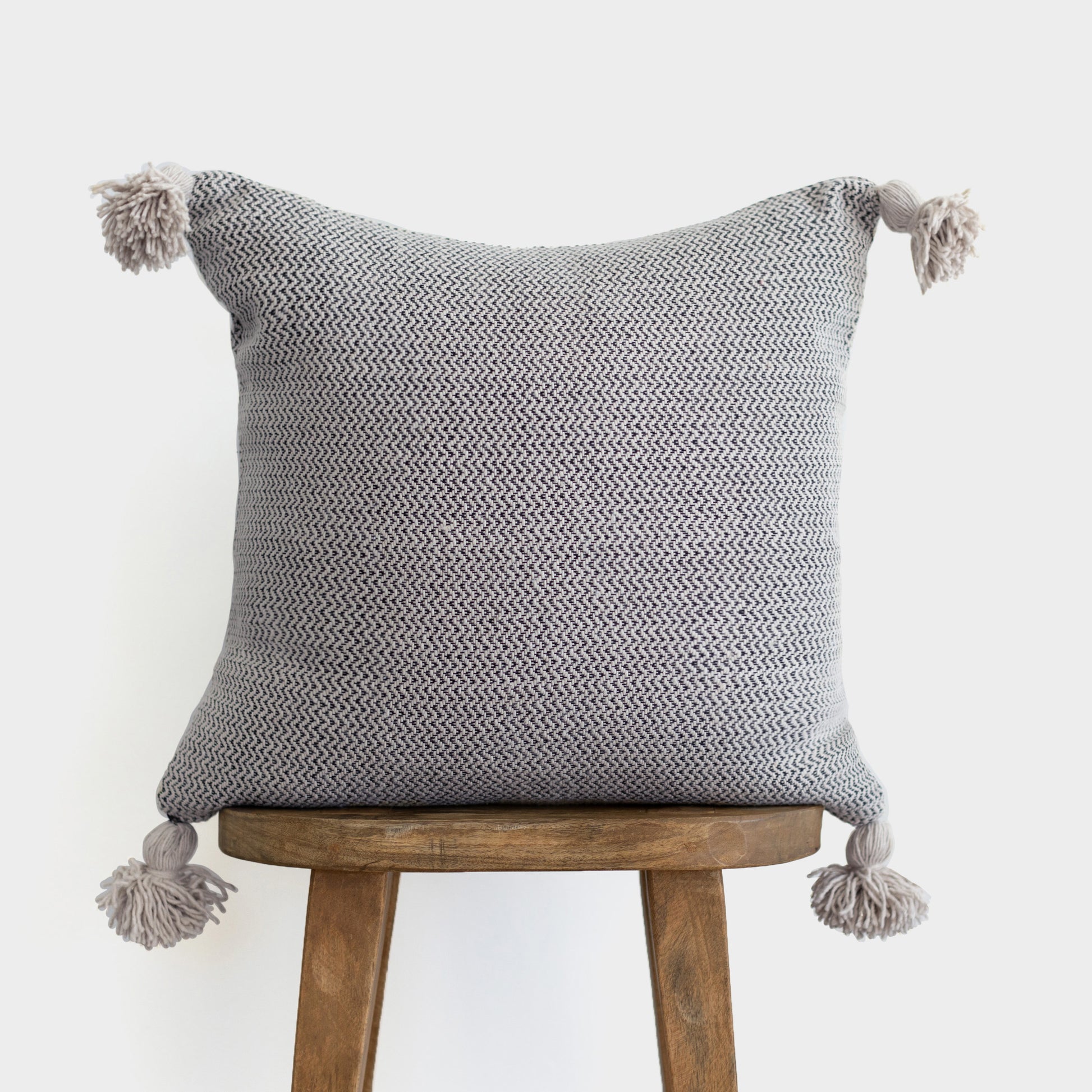 Gray tweed pillow with chevron pattern and tassel corners displayed on wooden stool.