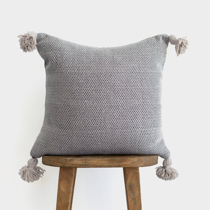 Gray tweed pillow with chevron pattern and tassel corners displayed on wooden stool.