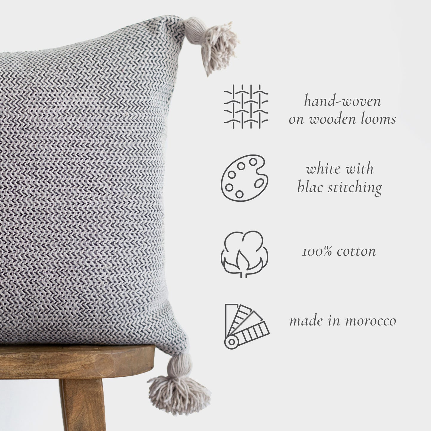 Gray and white hand-woven cotton pillow with black stitching and tassels on wooden chair.