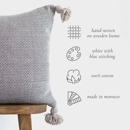 Gray and white hand-woven cotton pillow with black stitching and tassels on wooden chair.