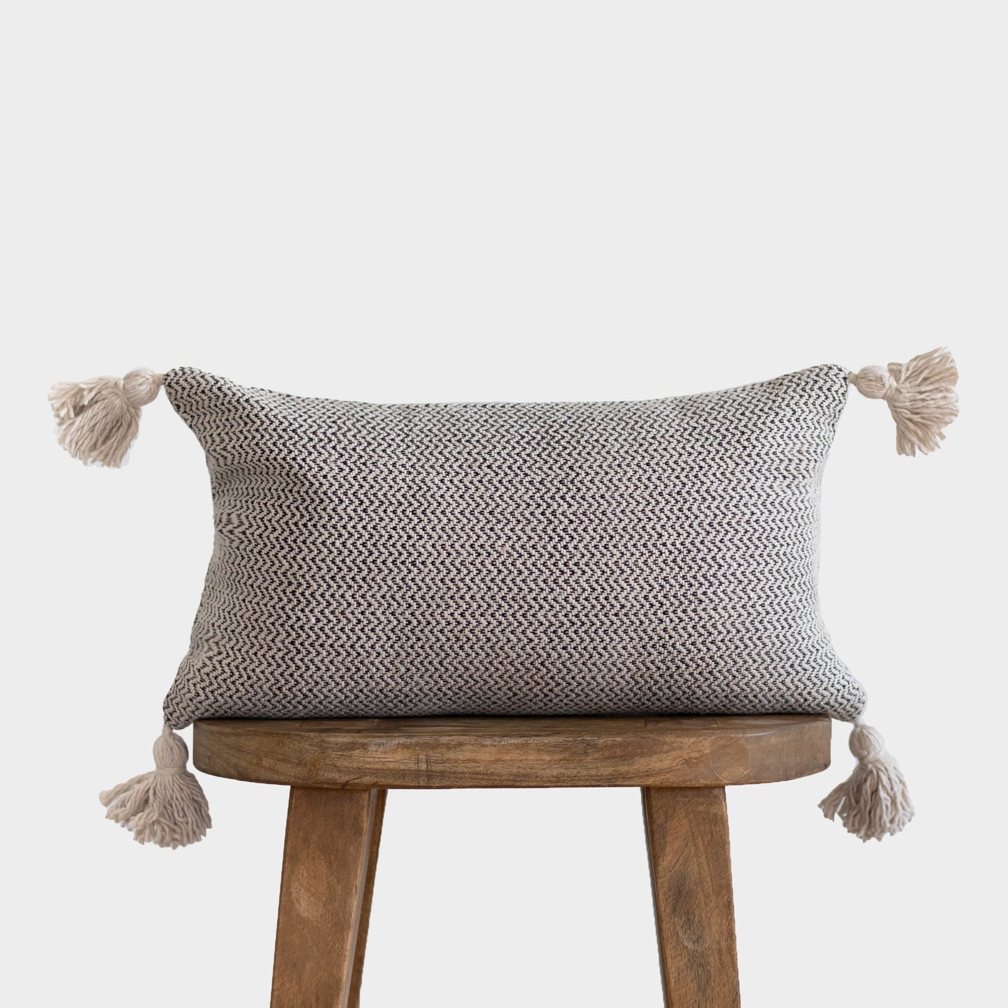 Cream and gray tweed rectangular pillow with tassels on four corners, displayed on wooden stool.