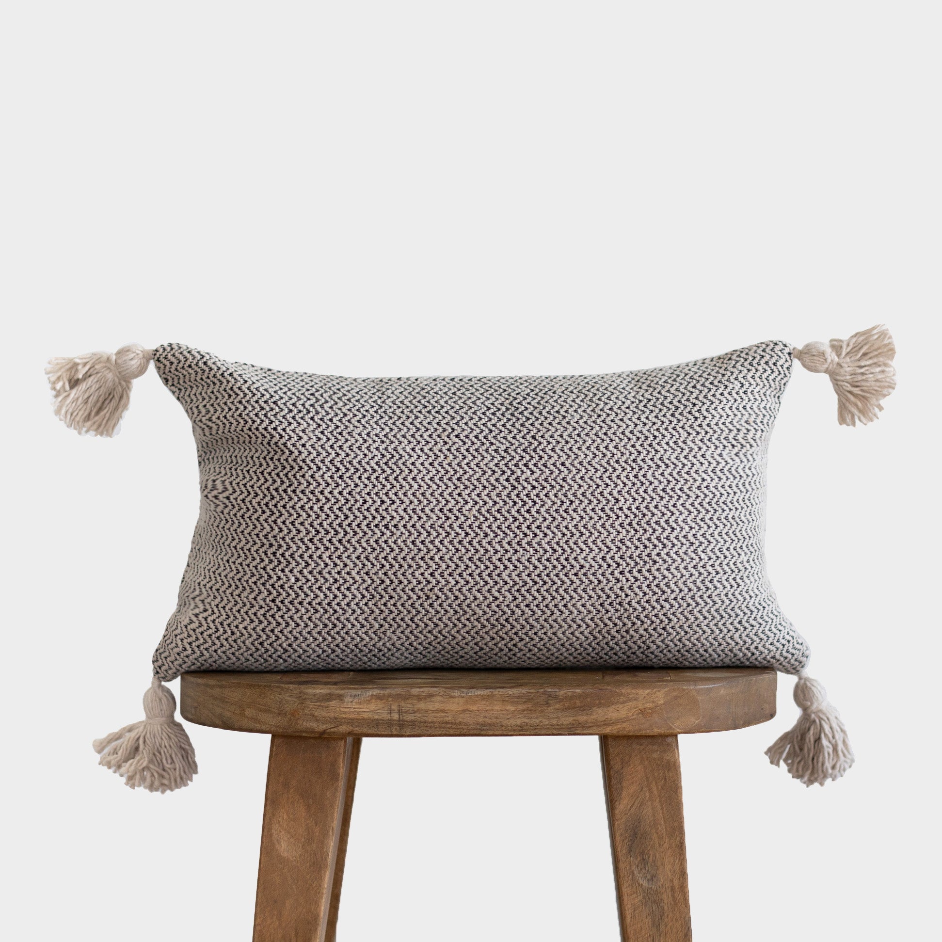 Cream and gray tweed rectangular pillow with tassels on four corners, displayed on wooden stool.