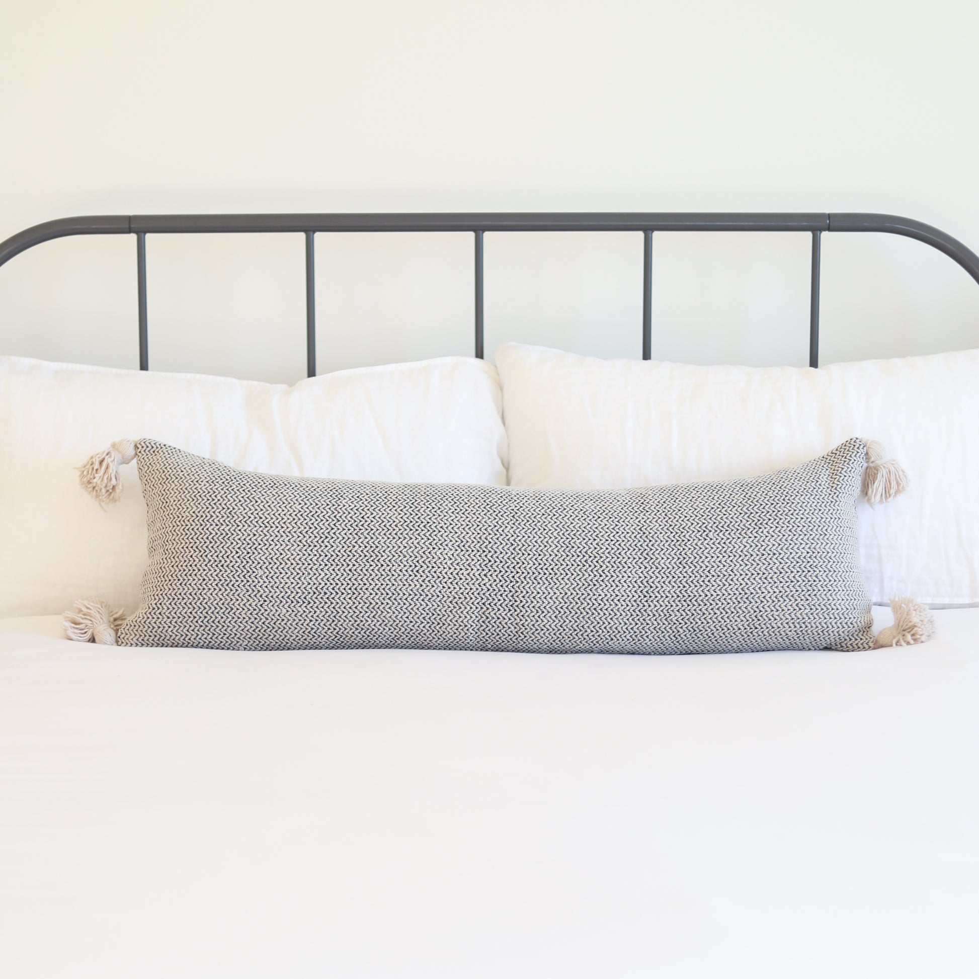 Cream tweed lumbar pillow with tassel corners displayed on a bed with white pillows.