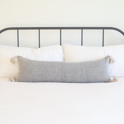 Cream tweed lumbar pillow with tassel corners displayed on a bed with white pillows.