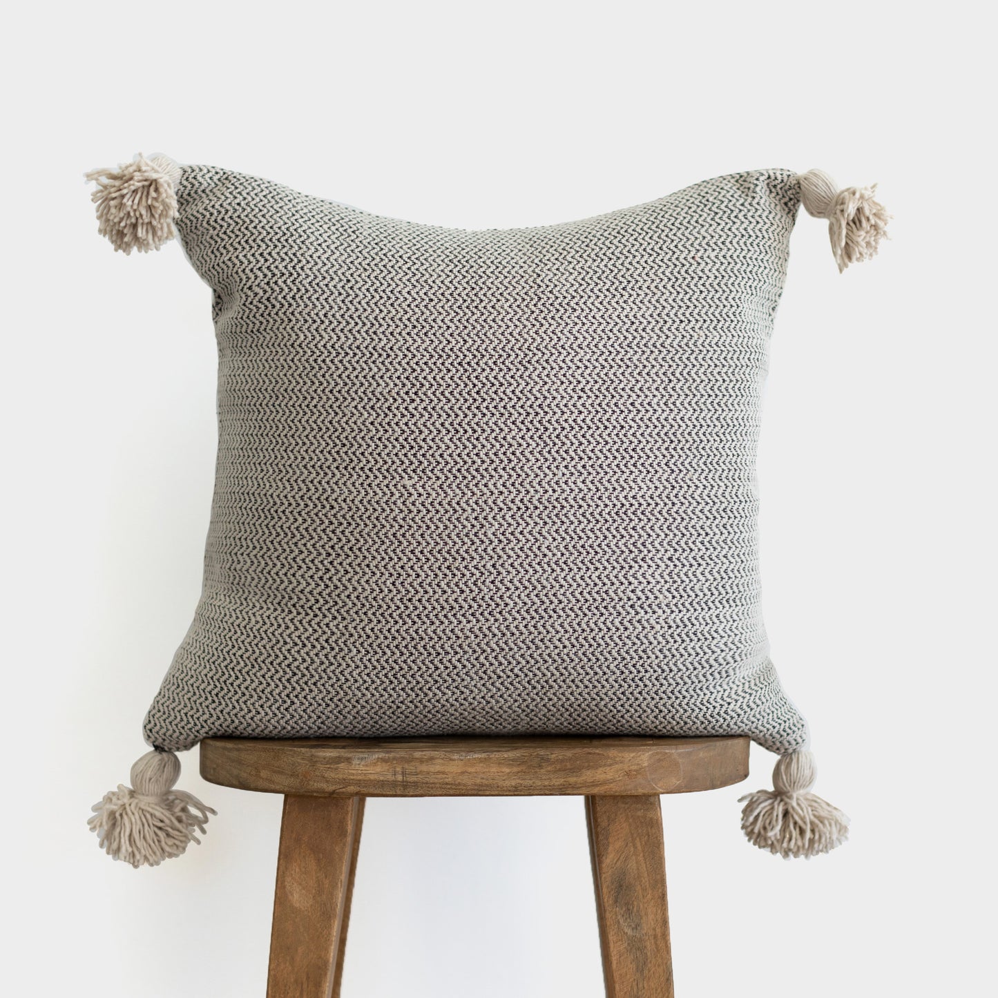 Cream and gray tweed pillow with corner tassels displayed on wooden stool.