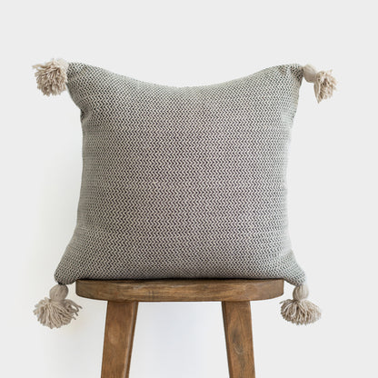 Cream and gray tweed pillow with corner tassels displayed on wooden stool.