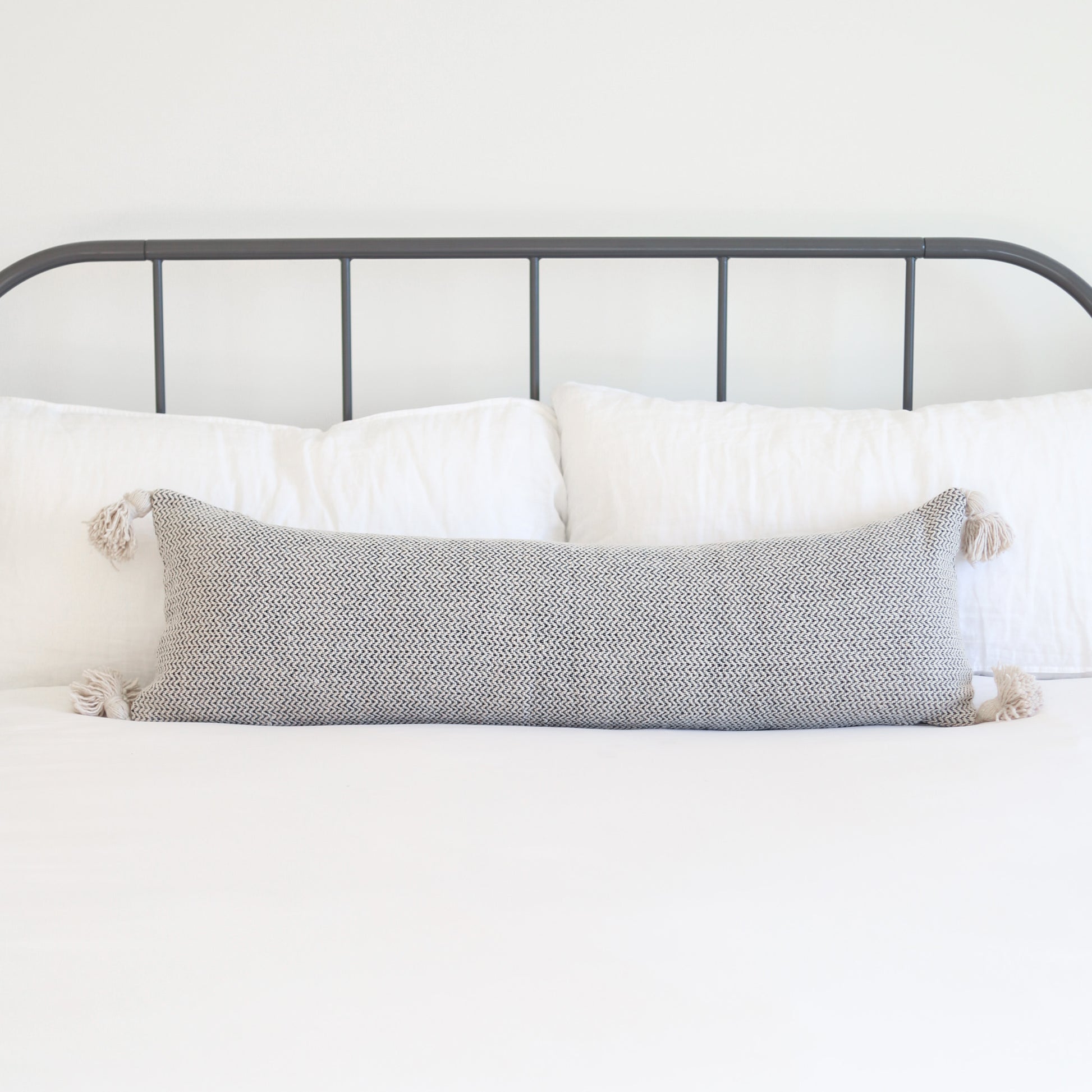 Gray tweed lumbar pillow with cream tassels on corners, displayed on bed with white pillows and metal headboard.